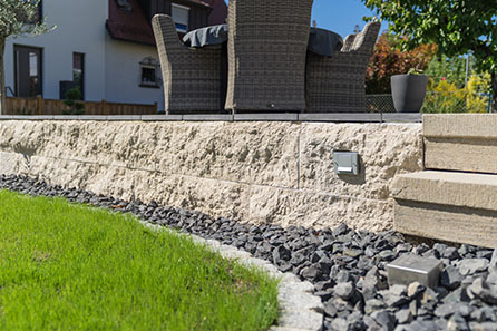 Runde Rasenkante mit Granitsteinen befestigt, dahiner eine kleine Schotterfläche vor einer erhöht liegenden Terrasse mit Steinbelag und geflochtenen Möbeln