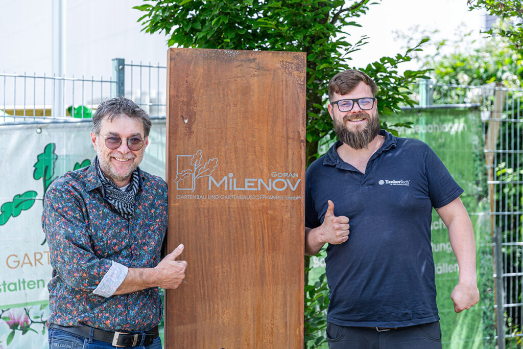 Goran Milenov und sein Mitarbeiter stehen vor einem Firmenschild in Rost-Optik mit nach oben gestreckten Daumen